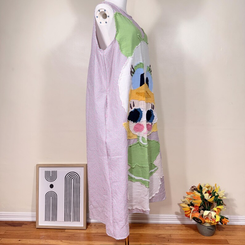 May include: A sleeveless, patchwork dress featuring a cartoon character design. The dress has a light purple and white checkered pattern on the left side, and a colorful cartoon face on the right side. The dress is displayed on a mannequin.