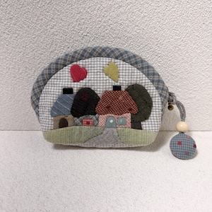 Può includere: Un portamonete trapuntato fatto a mano con una scena di villaggio. La borsa ha un motivo a quadri blu e grigio e una forma arrotondata. La scena del villaggio comprende case, alberi e cuori. Un cordoncino grigio con una perla di legno e una piccola pallina di stoffa completa il design.