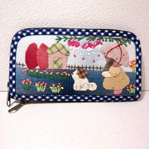 May include: A fabric wallet with a whimsical scene of a girl with an umbrella, a dog, and a house. The wallet has a blue and white gingham border and a zipper closure. The scene includes red trees, flowers, and a fence.