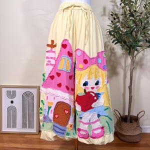 May include: A pair of pale yellow wide-leg pants featuring a drawstring waist and large, colorful fabric appliqués. The designs include a pink house with red hearts and a cartoon character with blonde hair holding a large red apple.