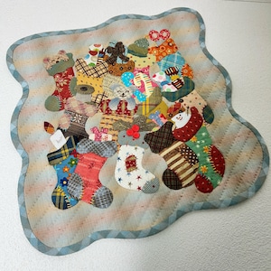 以下が含まれることがあります： 赤、緑、青、黄色の様々な生地で作られた、クリスマスツリーのデザインのカラフルなパッチワークキルト。ストッキング、オーナメント、その他のクリスマスの要素で構成されています。キルトは青と白のトリムで縁取られています。