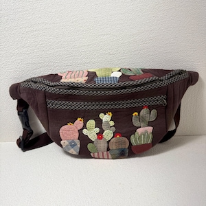 May include: A brown fanny pack with a zipper closure and a black buckle. The bag is decorated with colorful, appliqued cactus designs. The cacti are in various pots and have red and yellow flower accents.