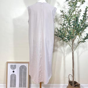 May include: A sleeveless, light beige dress with a round neckline and a slightly curved hemline. The dress is displayed on a mannequin. The background includes a framed art print and a potted plant.