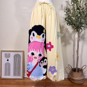 Könnte beinhalten: Hellgelbe Hose mit weitem Bein und elastischem Kordelzug in der Taille. Auf der Vorderseite befindet sich eine große, strukturierte Applikation eines Mädchens mit rosa Haaren, blauem Kleid und roter Tasche, flankiert von zwei schwarz-weißen Pinguinen. Weitere bunte Blumenapplikationen sind an der Seite verteilt.