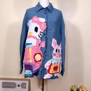 May include: A blue long-sleeved button-down shirt. The shirt is decorated with appliqued cartoon characters in pink, white, and yellow. The characters include a cat and a rabbit. The shirt is displayed on a mannequin.