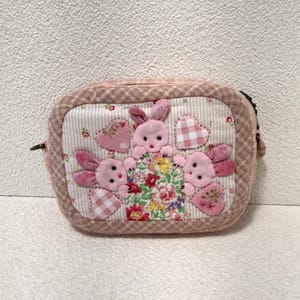 Puede incluir: Un estuche acolchado rectangular con fondo de rayas rosa claro y blanco, bordeado con un patrón de cuadros vichy rosa y blanco. El frente muestra tres conejos de tela rosa, detalles de tela floral y varios corazones de cuadros y flores.