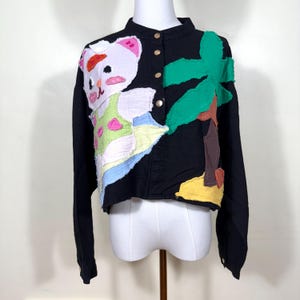 Può includere: Giacca nera con un design stravagante con un orso bianco con un cappello rosso e arancione, una palma verde e una spiaggia gialla. La giacca ha bottoni dorati e maniche lunghe. L'orso indossa un completo verde e rosa.