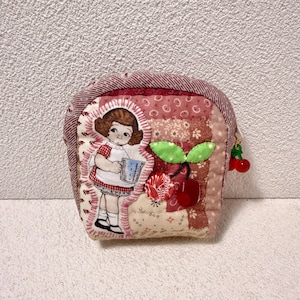Könnte beinhalten: Ein gestepptes Münzportemonnaie mit Patchwork-Design in Rosa- und Rottönen. Auf der Vorderseite befindet sich eine Illustration eines Mädchens, das einen blauen Krug hält, und eine Kirschapplikation. Ein kleiner Kirschanhänger hängt am Reißverschluss.