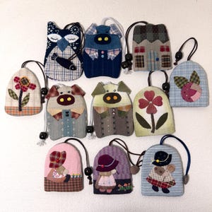 May include: A collection of handmade fabric keychains in various shapes and designs. The keychains feature animal and house motifs, including owls, pigs, and houses, with floral and character embellishments. Each keychain has a drawstring closure and a black loop.