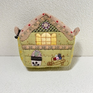 Pode incluir: Uma pequena bolsa de moedas de tecido em forma de casa verde e marrom com telhado rosa e vermelho, uma janela amarela e um design floral. A bolsa tem uma pequena alça de cada lado.