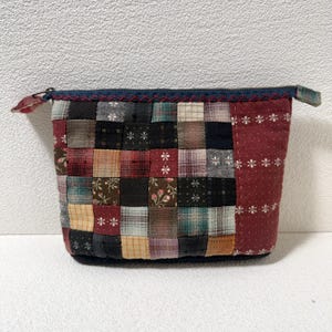 Puede incluir: Una bolsa con cremallera de tela de retazos con una variedad de cuadrados en tonos de rojo, marrón, negro y crema. La bolsa tiene una cremallera azul oscuro y un panel de tela roja con detalles florales blancos.