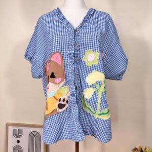 Può includere: Camicia a quadri blu e bianca con scollo a V e maniche corte. Disegni applicati di un orsetto dei cartoni animati e fiori decorano la parte anteriore. Il colletto e l'abbottonatura sono rifiniti con volant.