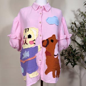 May include: A light pink button-down shirt with ruffled puff sleeves and a textured fabric. The front features large, colorful fabric appliques of a yellow cat wearing a purple outfit and a brown animal, plus a small light blue cloud patch.