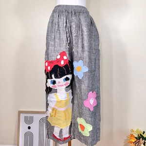 May include: Gray wide-leg pants with an elastic waistband. The pants feature an applique of a cartoon girl with a red bow and floral accents in blue, pink, and green. The girl is wearing a yellow dress.