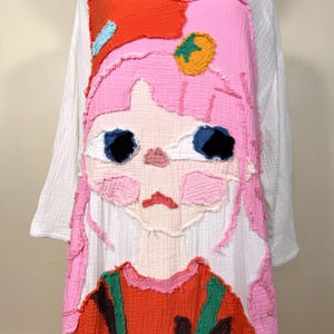 May include: A white long-sleeved dress with a large, colorful applique of a cartoon girl's face. The face features pink hair, blue eyes, and a red and orange outfit. The applique is made of textured fabric.