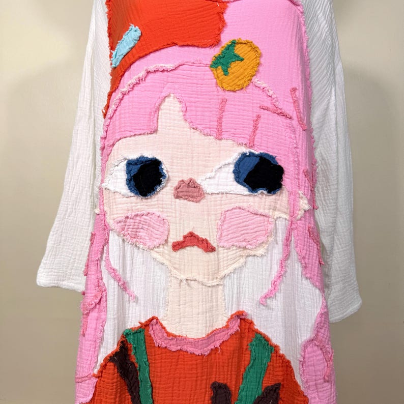 May include: A white long-sleeved dress with a large, colorful applique of a cartoon girl's face. The face features pink hair, blue eyes, and a red and orange outfit. The applique is made of textured fabric.