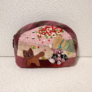 Puede incluir: Un estuche redondo hecho a mano con un diseño caprichoso. El estuche presenta un patchwork de telas en tonos rosa, marrón y verde. En la parte delantera se aplican un gato marrón, una niña con un vestido floral y adornos florales e insectos.