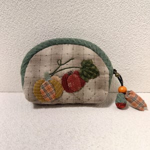 Puede incluir: Un pequeño monedero acolchado beige con ribete verde y una aplicación de calabaza y manzana. El bolso tiene un estampado de cuadros y una pequeña borla con una cuenta de madera. El diseño recuerda al otoño.