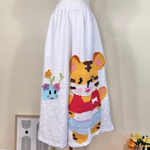 Może przedstawiać: Biała spódnica z teksturowanym wzorem, z dużą, kolorową aplikacją przedstawiającą tygrysa. Tygrys ma na sobie czerwoną koszulę i niebieskie szorty. Obecna jest również mniejsza aplikacja uśmiechniętej doniczki.