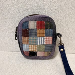 Pode incluir: Uma pequena bolsa quadrada com fecho de correr, apresentando um painel frontal de patchwork composto por vários quadrados de tecido multicoloridos com padrões xadrez, listrados e lisos. A bolsa tem uma borda roxa escura e uma alça de pulso azul.