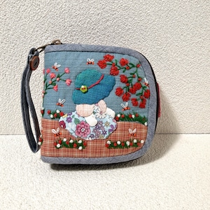 May include: A blue fabric zippered pouch with a wrist strap featuring embroidered details of a figure in a teal hat sitting in a garden with red flowers, green foliage, and small bees. The lower section has a brown and orange plaid pattern.