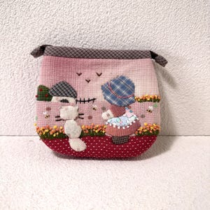 May include: A small fabric pouch featuring a quilted scene of a person in a blue plaid bonnet and a white cat in a garden. The design includes embroidered flowers, bees, a house, and birds against a pink background with a red polka dot base.