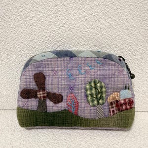 May include: A handmade, lavender-colored, zippered pouch with a whimsical landscape design. The pouch features a brown windmill, trees, and a house, all crafted from various fabrics. The top is patterned with blue and gray diamonds.