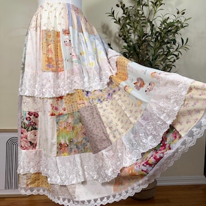 May include: A tiered patchwork skirt featuring a variety of floral, plaid, and cartoon-patterned fabric panels in pastel and muted tones. The skirt includes multiple horizontal layers accented with white lace trim and an elasticized waistband.