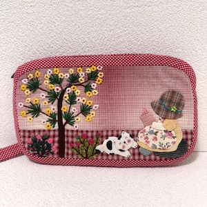May include: A rectangular, quilted pouch with a red and white polka dot border. The front features an embroidered scene of a tree with yellow flowers, a girl in a floral dress, a cat, and a checkered background.