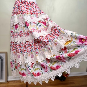 Op de afbeelding: Een gelaagde rok met bloemenprint en witte kanten rand. De rok heeft een verscheidenheid aan kleurrijke bloemen, waaronder rood, roze en geel, op een witte achtergrond. De rok wordt op een mannequin gepresenteerd.