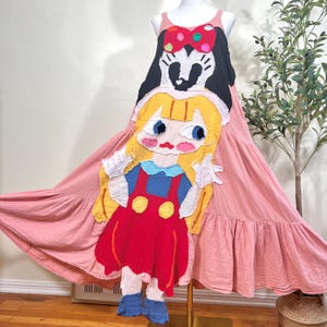 May include: A pink, sleeveless dress with a tiered skirt. The dress features a large, colorful appliqué of a cartoon character. The appliqué includes a black head with a red bow, and a yellow-haired character in a red dress.