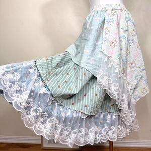 May include: A layered skirt with a whimsical design. The skirt features multiple tiers of fabric in pastel colors, including light blue, green, and white. The top layer has a cartoon character print, while the middle layer has a striped pattern with small flowers. The bottom layer has a plaid pattern with white lace trim.