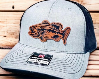 May include: Gray and black trucker hat with a brown leather bass fish patch. The hat has an adjustable strap and the text "Richardson 112 Trucker Adjustable" on the back.