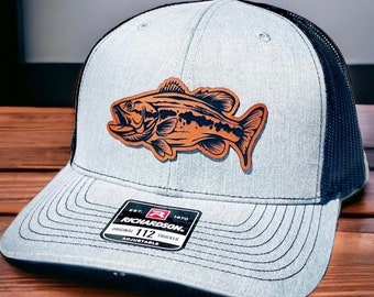 May include: A gray and black trucker hat with a brown leather bass fish patch. The hat has a black mesh back and a black brim. The hat has a black adjustable strap with a black buckle. The hat has a black and red label that says "Richardson 112 Original Trucker Adjustable".