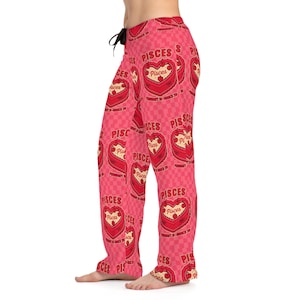 Puede incluir: Pantalones de pijama rosas con un patrón repetido de pasteles en forma de corazón y la palabra "Pisces". Los pantalones tienen una cintura con cordón y un fondo a cuadros. El diseño de Piscis es en rojo y blanco.