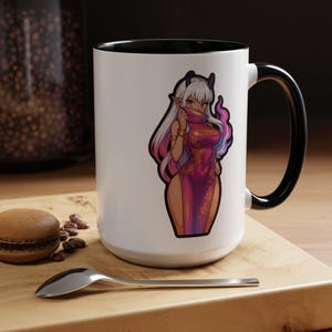 May include: White ceramic mug with a black interior and handle. The mug features a colorful illustration of a character in a pink dress. A spoon, a macaron, and coffee beans are on a wooden surface.