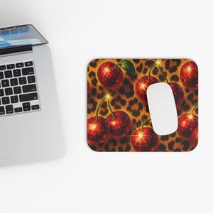 May include: A rectangular mousepad featuring a pattern of red disco ball cherries with green leaves against a leopard print background. A white computer mouse rests on the pad, next to a laptop.