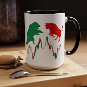 May include: White ceramic mug with a black interior and handle. The mug features a green bear and a red bull, with a stock market graph design. A perfect gift for investors and finance enthusiasts.