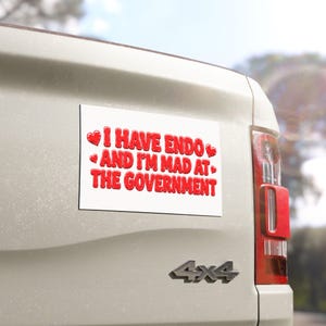 May include: A rectangular white bumper sticker with red text that reads I HAVE ENDO AND I'M MAD AT THE GOVERNMENT, accented by four small red heart icons. The sticker is adhered to the tailgate of a light-colored vehicle with a 4x4 emblem.