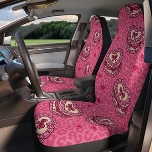 Puede incluir: Fundas de asiento de coche rosas con un patrón repetido de corazones y estampado de leopardo. Cada funda de asiento presenta un diseño de corazón grande con un lazo y un ribete de encaje. El interior de un coche es visible en el fondo.