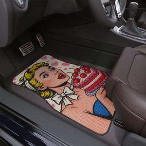 May include: Car floor mat featuring a pop art style illustration of a woman holding a cherry cake. The mat has a pink background with heart shapes and a black border. The woman has blonde hair and blue eyes.