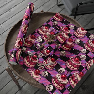 May include: A roll of patterned wrapping paper and a wrapped gift featuring a repeating design of pink cakes with cherries, dice, and black and purple checkered backgrounds. Scissors and a bow are also visible.
