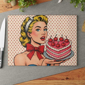 May include: A rectangular cutting board featuring a retro pop art illustration of a woman holding a heart-shaped cake topped with cherries. The woman has blonde hair, blue eyes, and a red bow. The background has a red polka dot pattern.