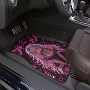 May include: A rectangular car floor mat featuring a vibrant pink and black graphic illustration of an anime-style character with long light hair. The design includes Japanese text, a crown icon, and heart symbols printed on a dark, textured background.