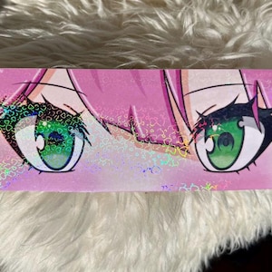 Puede incluir: Una pegatina rectangular que muestra un primer plano de grandes ojos de estilo anime con iris de color verde vibrante y pestañas oscuras. El fondo es de un rosa suave y toda la superficie tiene un acabado holográfico que refleja la luz.
