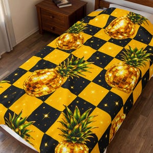 Könnte beinhalten: Eine gelb-schwarz karierte Decke mit einem sich wiederholenden Muster aus goldenen Ananas-Designs. Die Ananas haben eine mosaikartige Textur. Die Decke liegt auf einem Bett und wirkt einladend.