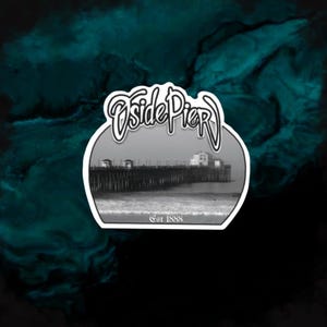 Könnte beinhalten: Ein Aufkleber mit einem Schwarz-Weiß-Bild eines Piers, der sich ins Meer erstreckt. Der Aufkleber trägt die Worte "Oside Pier" in einer stilisierten Schriftart, mit "Est 1888" darunter, vor einem teal-schwarzen Hintergrund.