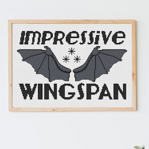 May include: A framed cross-stitch artwork with the words "IMPRESSIVE WINGSPAN" in black block letters. Two gray bat wings are centered below the word "IMPRESSIVE", with three small stars between them. The frame is a light wood color.