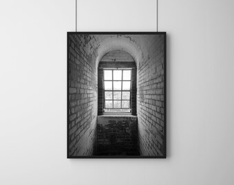 May include: Black and white photograph of a brick wall with a window. The window is arched and has a view of trees outside. The window is in a dark hallway.