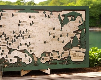 Puede incluir: Un mapa de madera de los Estados Unidos con un borde verde y un diseño de árboles recortados. El mapa está montado en un soporte de madera y tiene pequeños árboles de madera colocados en él para marcar ubicaciones. El texto "Ian & Krista's Adventure Map" está en la esquina inferior derecha del mapa.